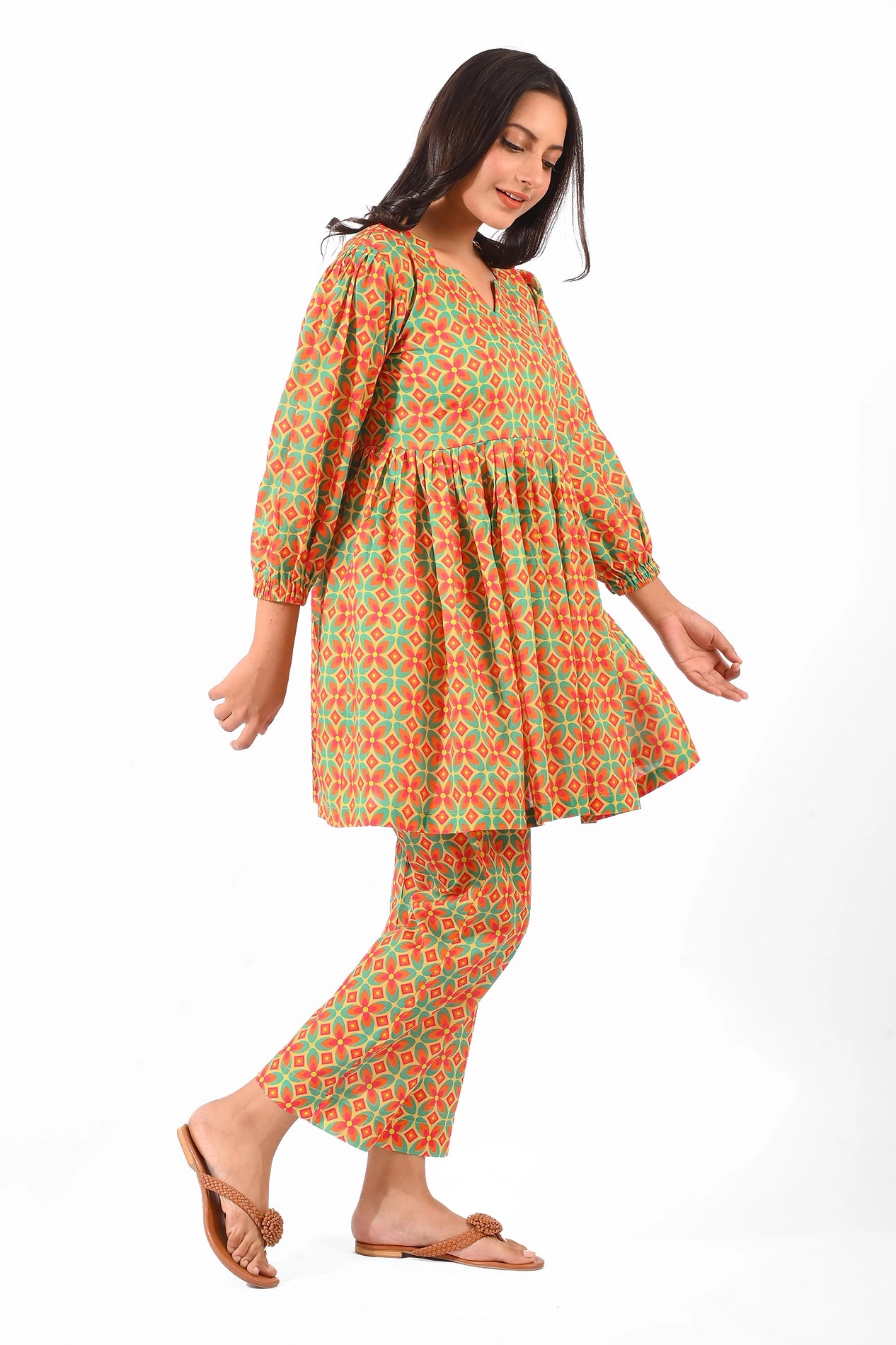 A side profile of a model wearing a colorful orange and green patterned dress with puffed sleeves and matching straight pants. The outfit has a gathered waistline and is paired with flat sandals, showcasing a casual and flowy design BOLDSTEP BOLDSTEP.PK