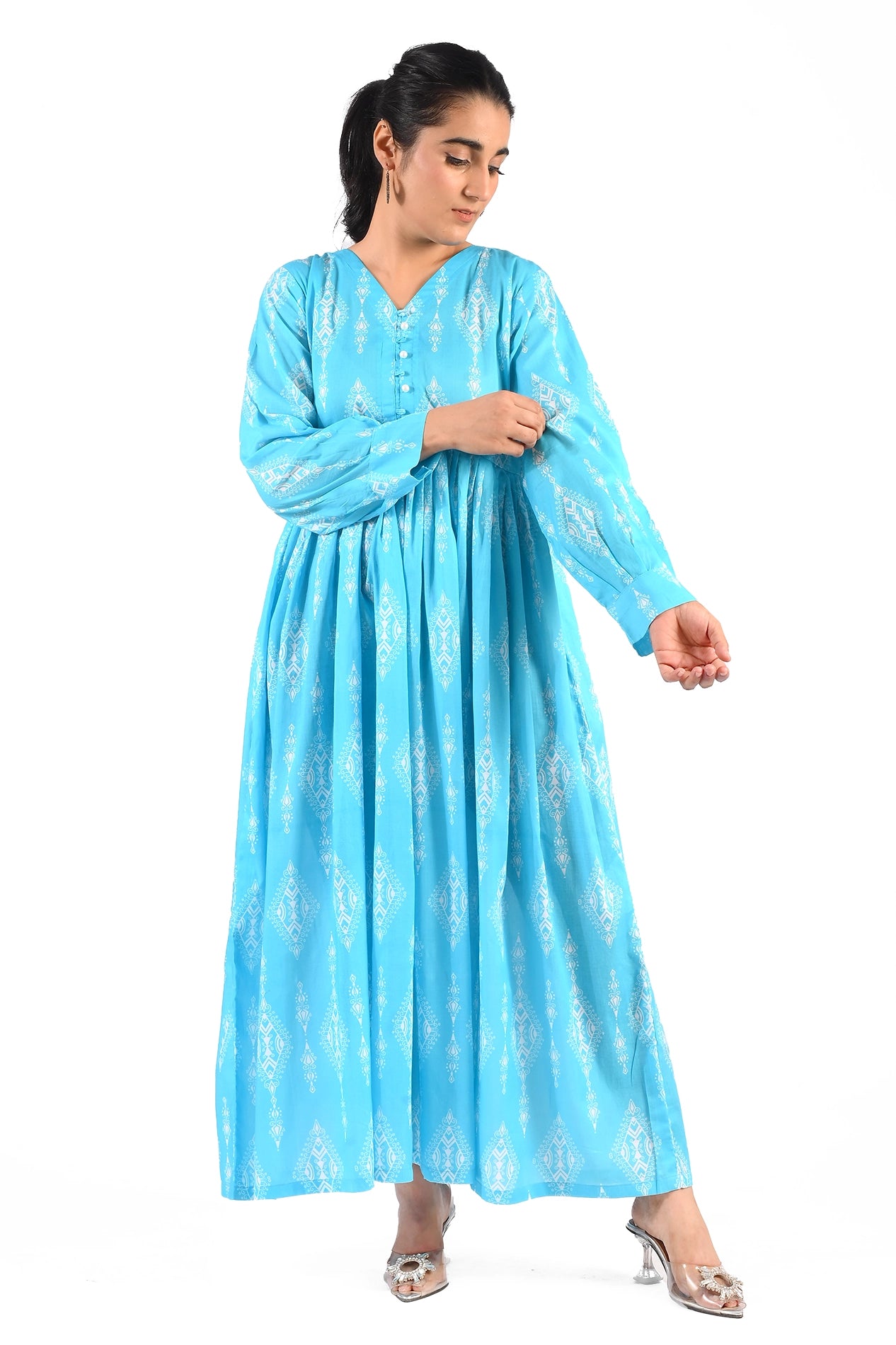A woman is wearing a blue maxi dress with a white geometric print. The dress has long sleeves, a V-neckline, and a button detailing on the bodice. She is posing with her hands adjusting her sleeve. BoldSTep BoldStep.pk