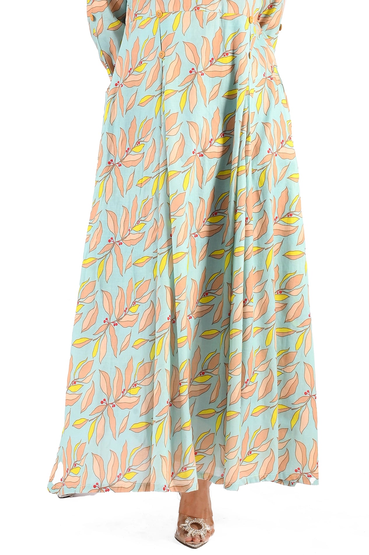A full-length maxi dress in a light aqua blue base adorned with soft peach and yellow leaf prints, accented by small red berries. The model stands facing forward, showcasing the dress's flowy design and button details. bOLDSTEP BOLDSTEP.PK