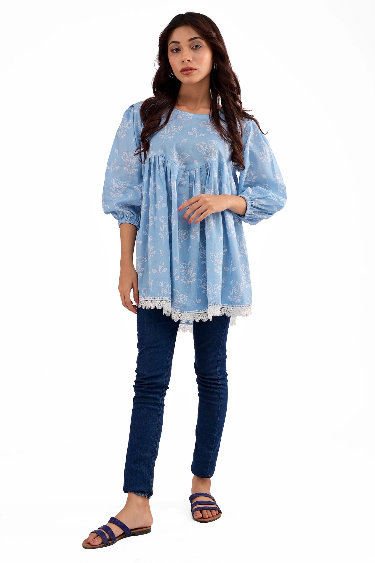 A model wearing a baby blue floral-printed tunic with a gathered empire waist, puffed long sleeves with elastic cuffs, and white lace detailing at the hem, paired with blue jeans and flat sandals