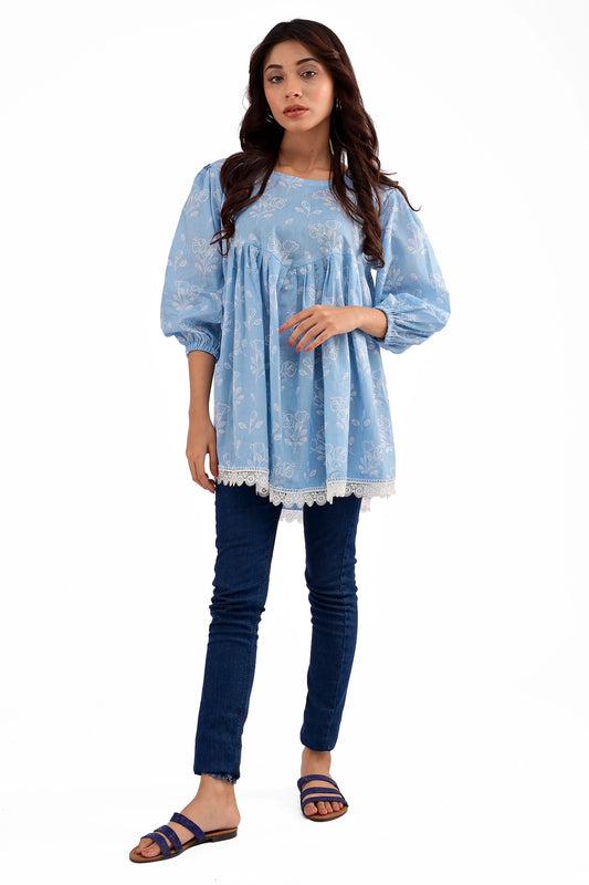 A model wearing a baby blue floral-printed tunic with a gathered empire waist, puffed long sleeves with elastic cuffs, and white lace detailing at the hem, paired with blue jeans and flat sandals