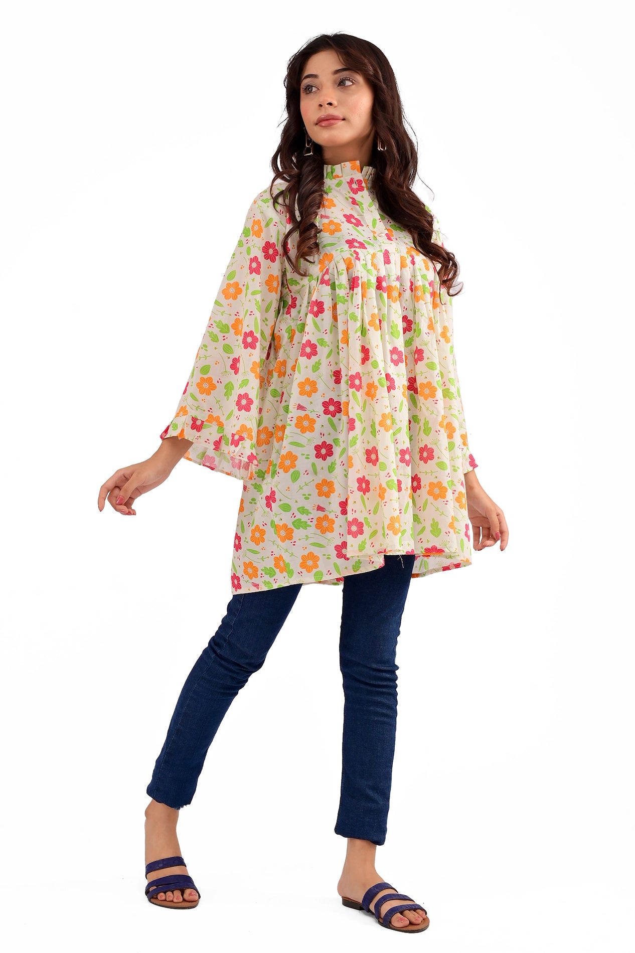 A model wearing a cream floral-printed tunic with bell sleeves and a high-neck buttoned collar, paired with dark blue skinny jeans and blue sandals