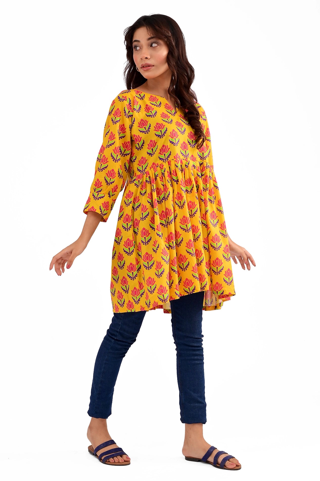 A model wearing a vibrant yellow tunic with a red and blue floral print, featuring a loose fit and three-quarter sleeves, paired with dark blue jeans and blue sandals