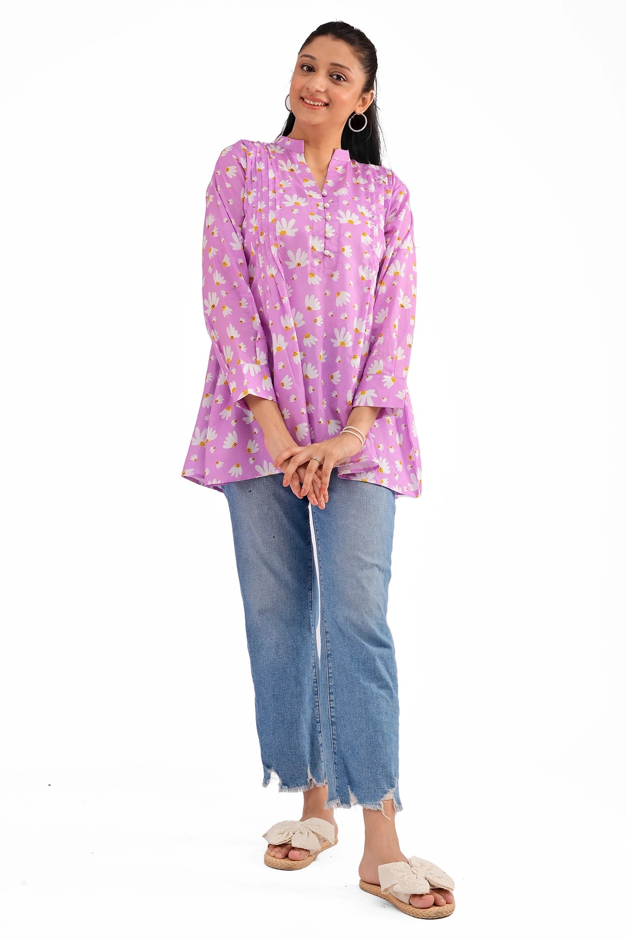 A model wearing a lavender tunic with a white daisy print, featuring a buttoned neckline and pleated front detail, paired with blue denim jeans and cream-colored sandals