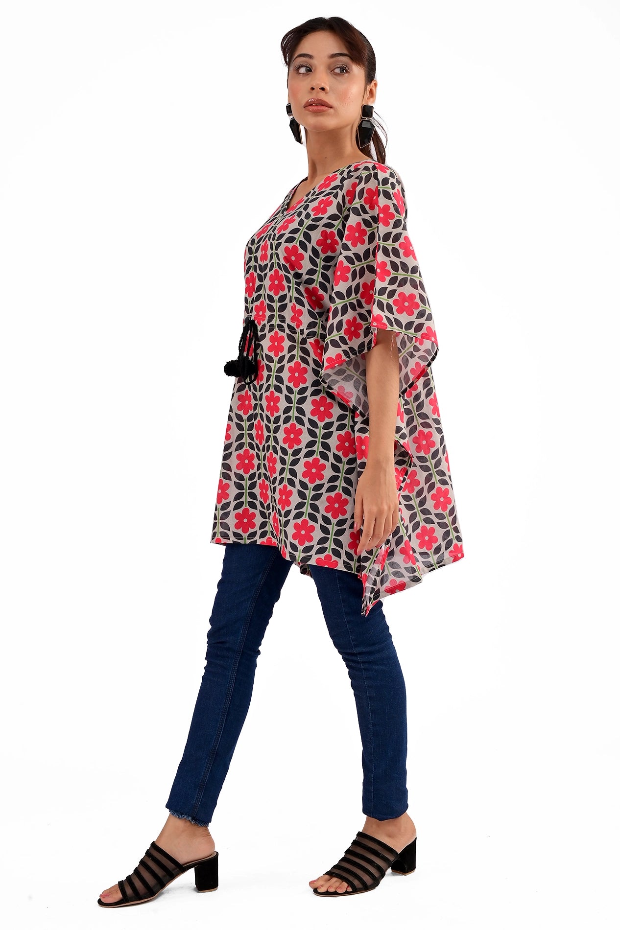 A side profile of a model wearing a black and red floral printed kaftan tunic with a V-neck and drawstring waist, styled with blue jeans and black heels