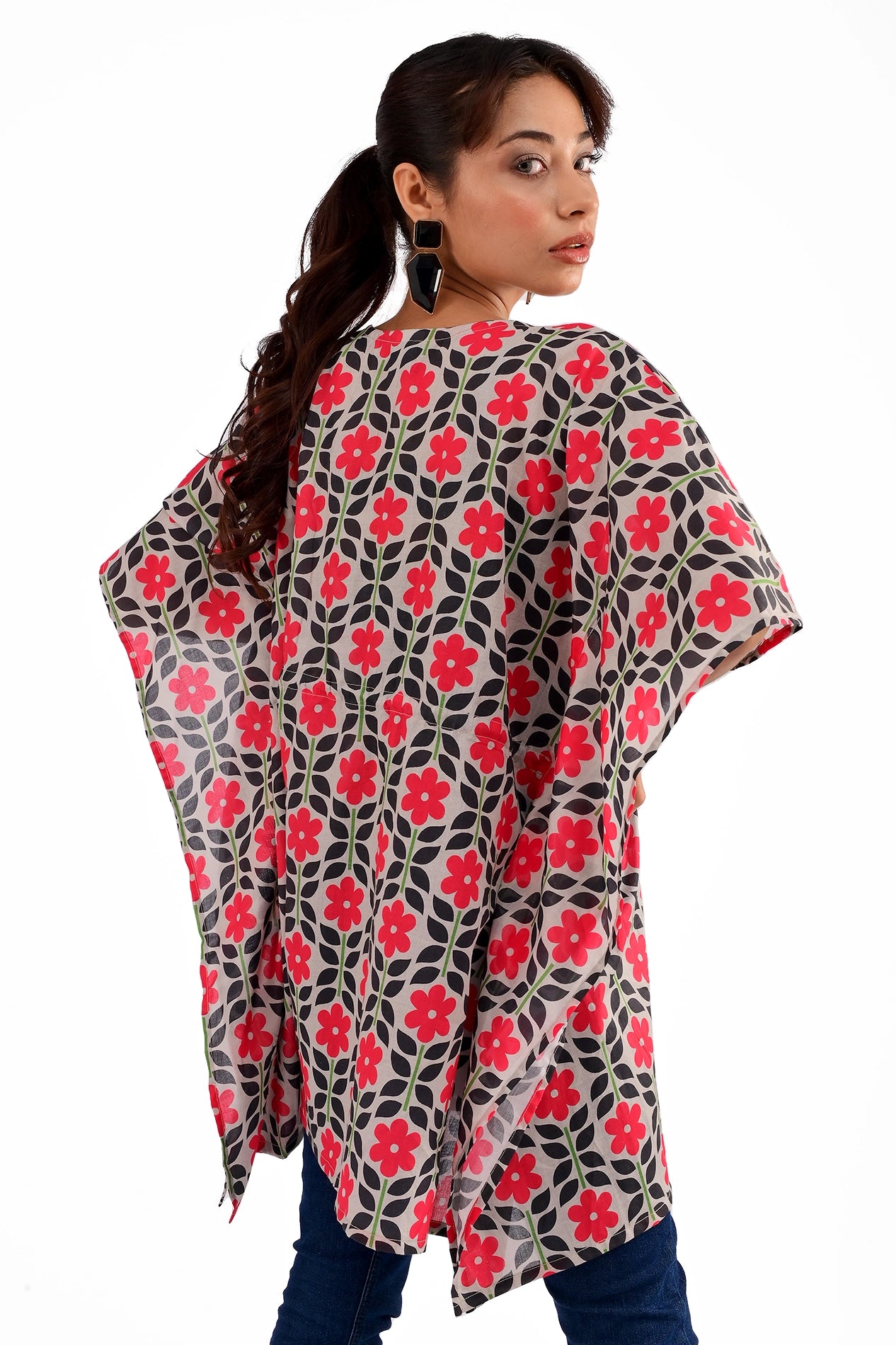 A rear view of a model wearing a floral printed black and red kaftan tunic with a relaxed fit, paired with blue jeans and black heels