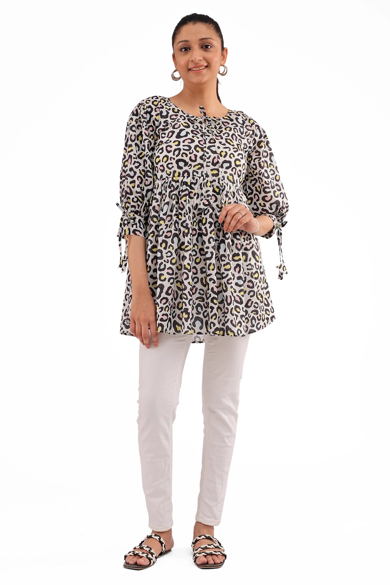 A model wearing a stylish leopard print tunic with tie-sleeves, paired with white slim-fit pants and braided sandals. The outfit has a relaxed fit with a gathered waist, offering a chic yet comfortable look