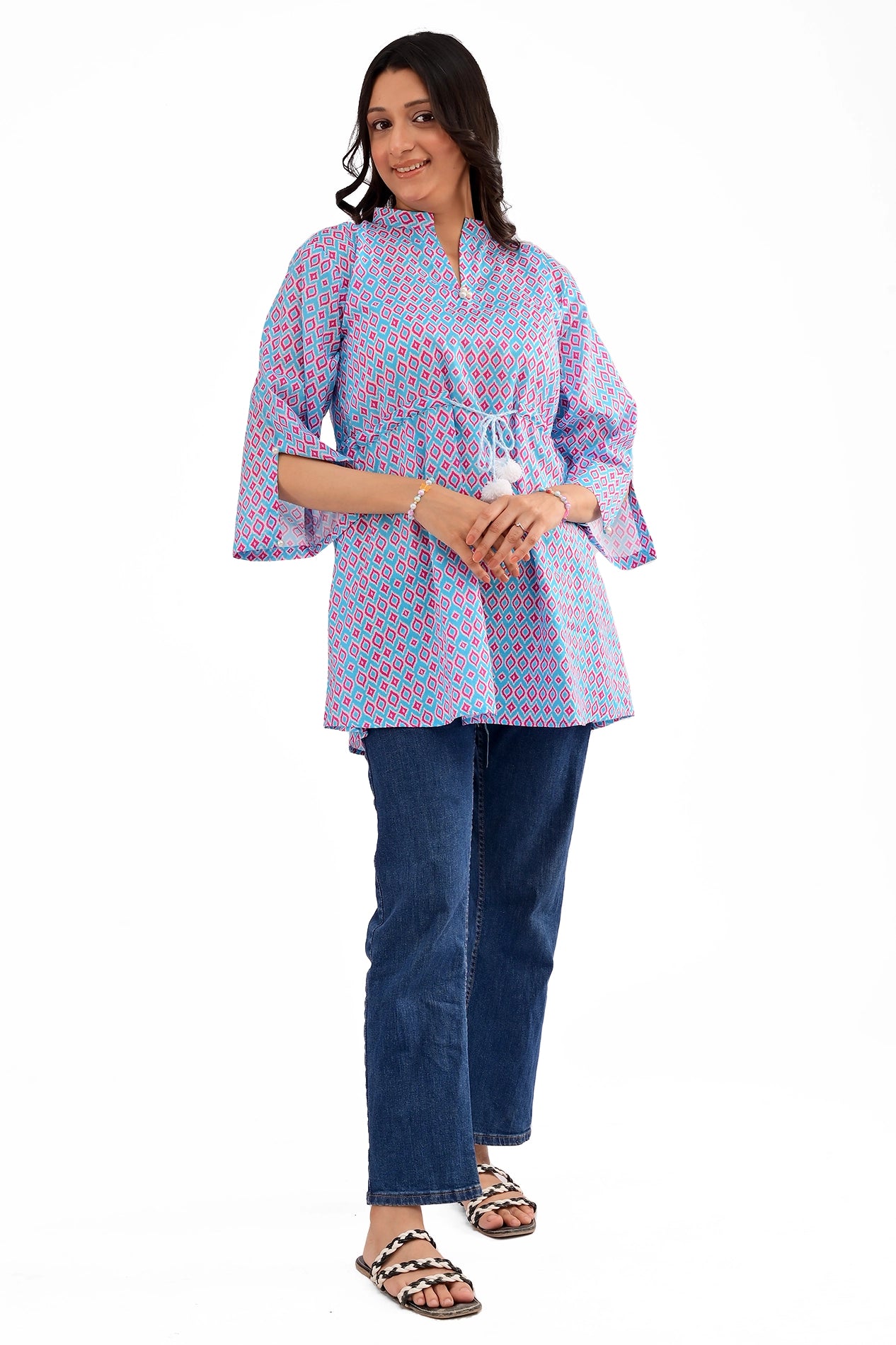 Woman wearing a blue and pink geometric print tunic with wide sleeves and a drawstring waist, paired with blue jeans and sandals