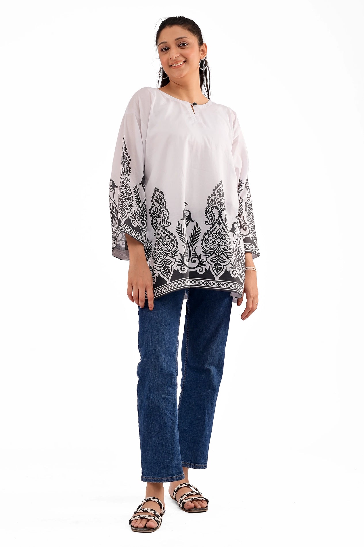 A woman wearing a white lawn kurti with intricate black traditional motifs along the hem and sleeves, paired with blue denim jeans and black sandals, posing with a smile.