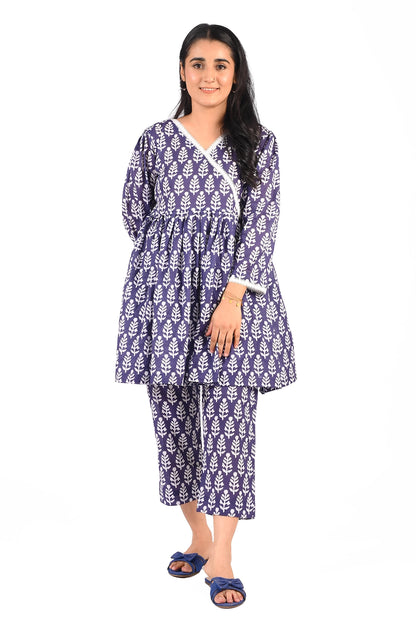 A smiling woman is dressed in a navy blue printed outfit featuring white leaf motifs. The ensemble consists of a knee-length wrap dress with a cinched waist and matching cropped trousers. She pairs the look with blue bow-detail flats BOLDSTEP BOLDSTEP.PK